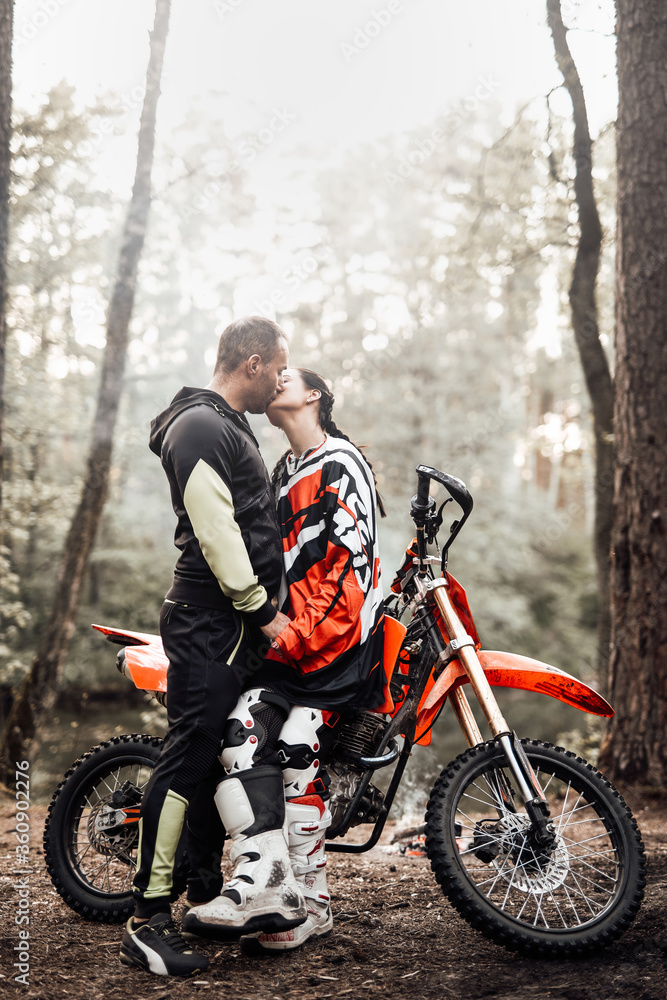Attractive couple kissing standing next to a motocross bike and bonfire in the woods