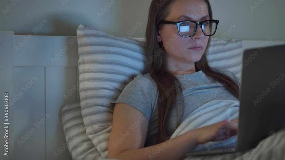 Woman in glasses working on laptop while lying in bed at night. Internet addict or insomnia concept. Woman in glasses working on laptop while lying in bed at night. Internet addict or insomnia concept.