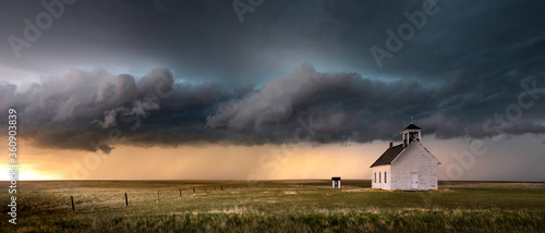 Old church in the rural countryside with a sever storm at sunset. There is an outhouse visible in the scene as well as a green and yellow grass meadow.