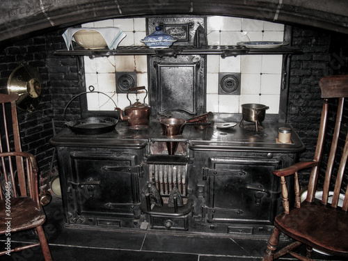 Old cast iron kitchen cooker, large stove with  ovens, grills , copper pots , pans , kettle, and other kitchen utensils, with ceramic tiles inset , and hint of wooden kitchen chairs just in view   