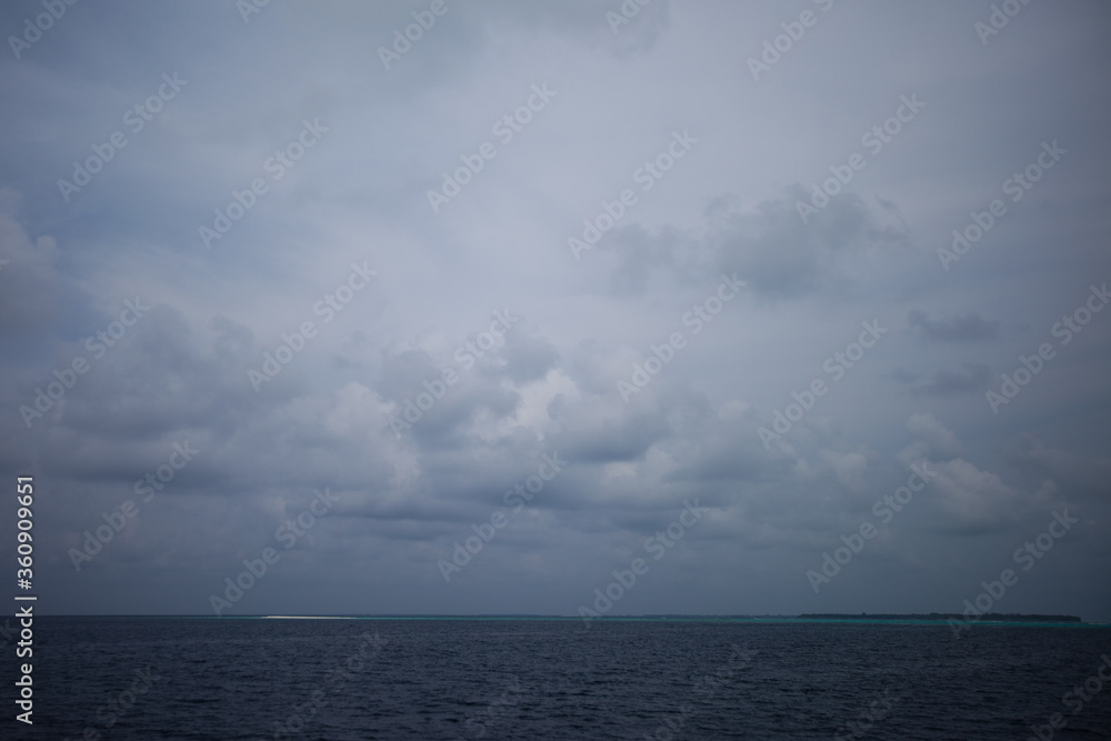 Cloudy day in Maldivian ocean with blue grey sky and blue grey water and white send island on the horizont