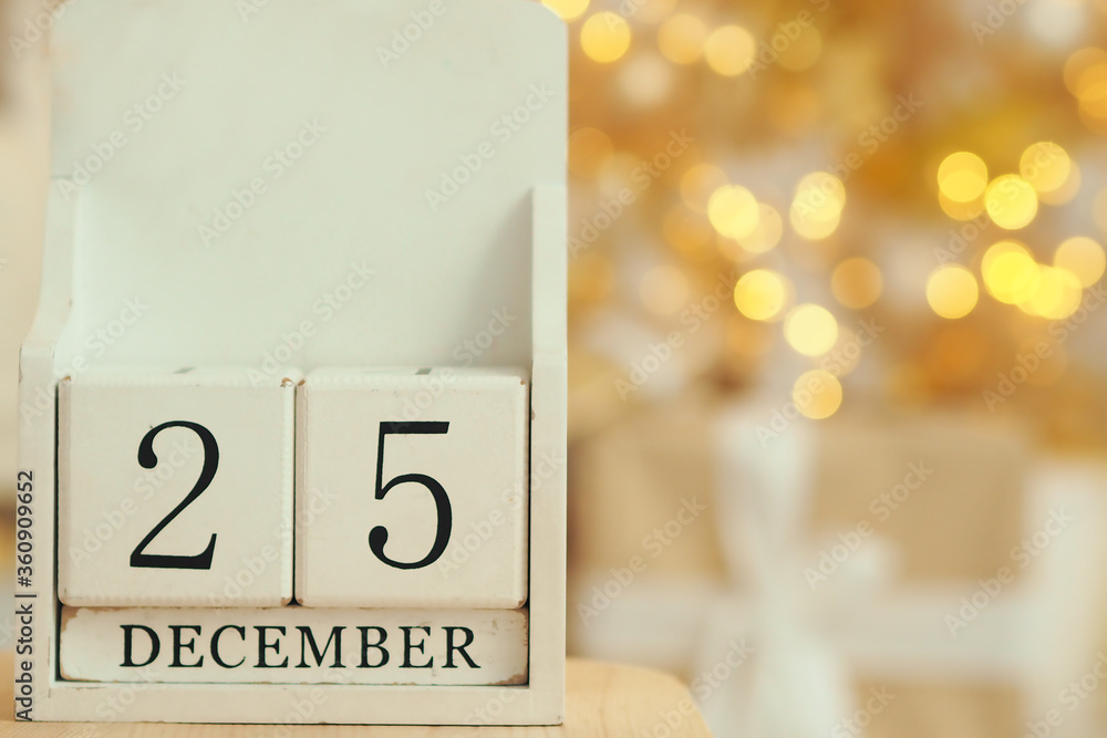 .White wooden calendar with cubes and the date December 25 and lights bokeh from a garland on a Christmas tree in the background. catholic christmas date. Copy space..