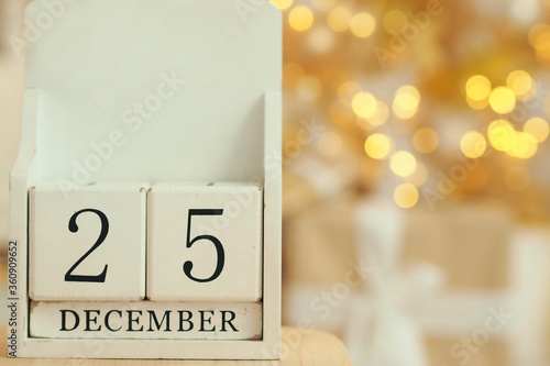 .White wooden calendar with cubes and the date December 25 and lights bokeh from a garland on a Christmas tree in the background. catholic christmas date. Copy space..