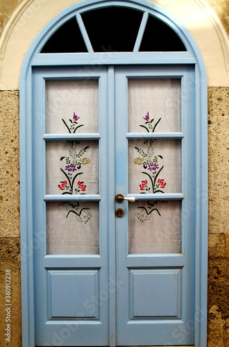 A celestial blue wooden door with floral pattern  embroidered curtains in the Aegean town Alacati, in Izmir, Turkey. 