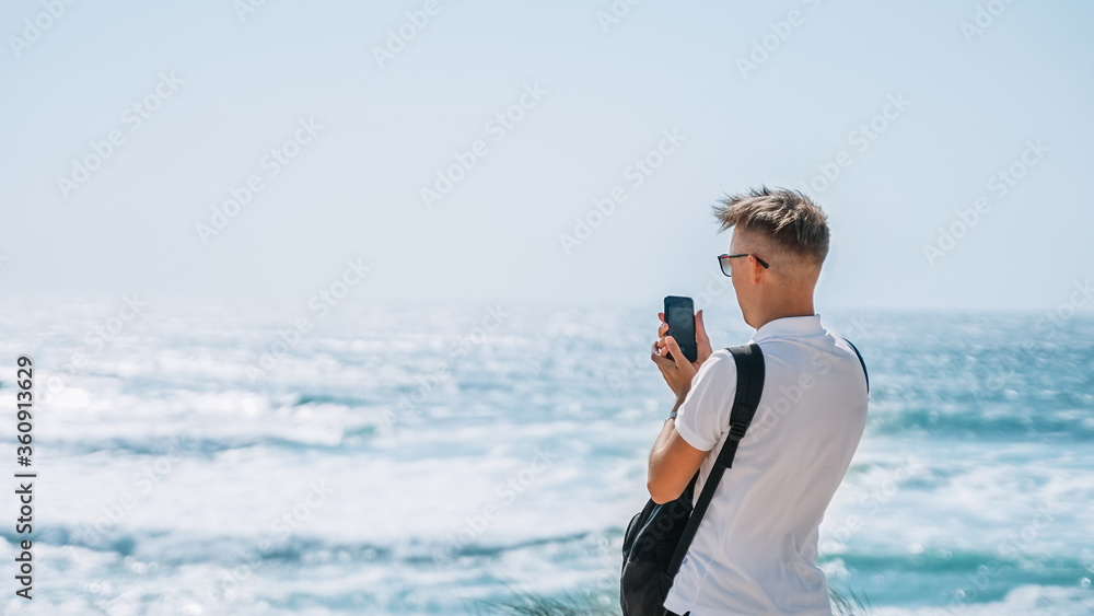 young man tourist taking a picture of the sea or ocean landscape. back view portrait. blue sky, horizon, ocean. traveler making photo recording video of vacation adventure exploring. wide banner
