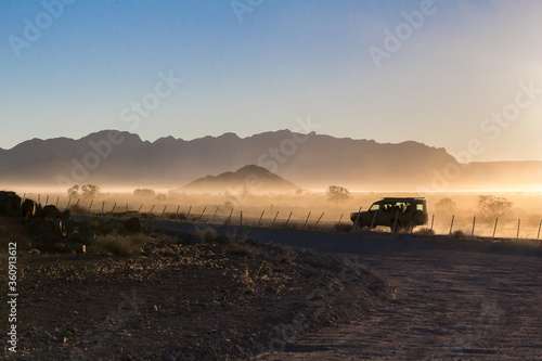 Coche jeep avanzando por una carretera de gravilla al amanecer. El vehículo está saliendo del Parque Nacional Namib-Naukluft por Sesriem, en Namibia.