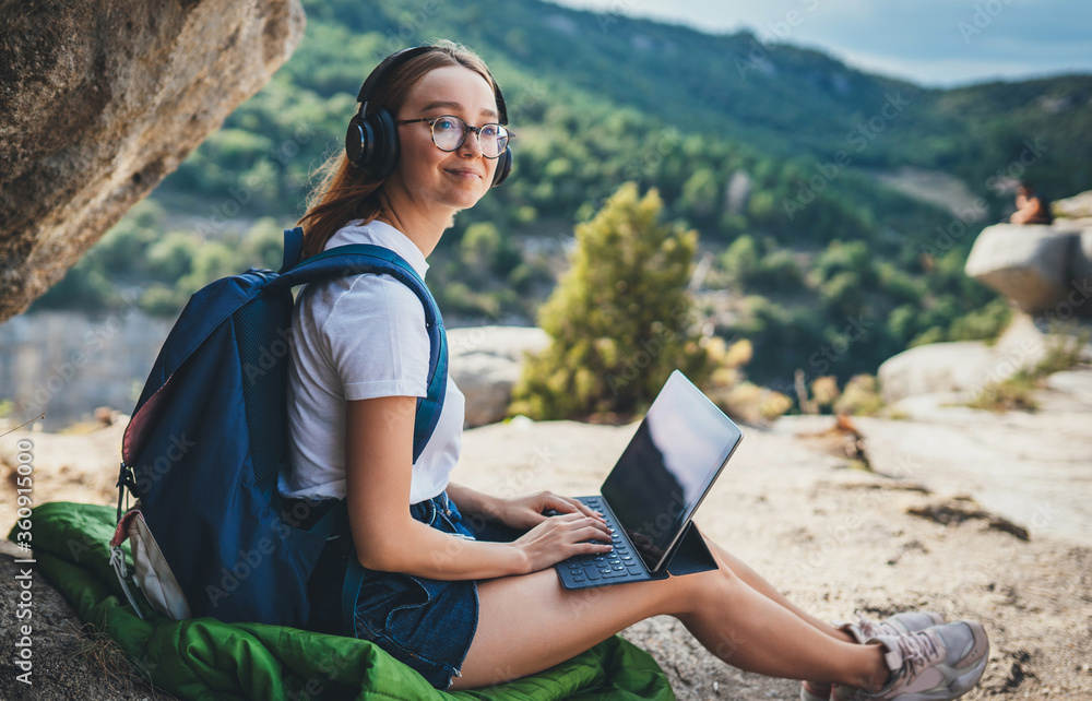 tourist girl with glasses and headphones using digital tablet device sitting in nature outdoor, hipster woman with backpack listen music headphones and laptop computer on backdrop of high mountains