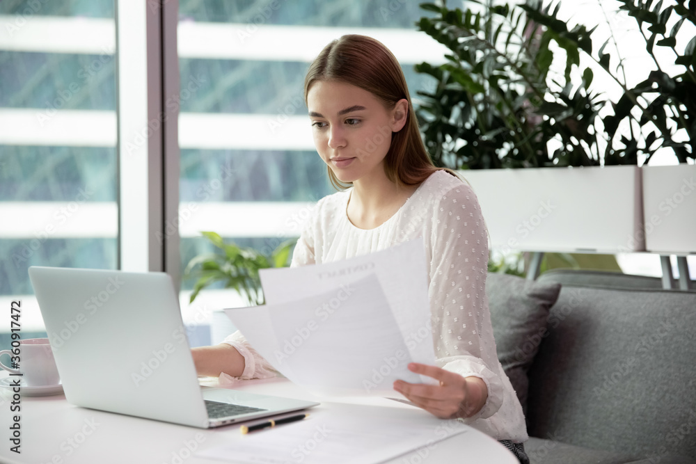 Focused millennial one female worker busy at laptop in modern office room. Woman manager or ceo sit and hold paper sheet or document use computer business networking. Businesswoman at desk read report Focused millennial one female worker busy at laptop in modern office room. Woman manager or ceo sit and hold paper sheet or document use computer business networking. Businesswoman at desk read report