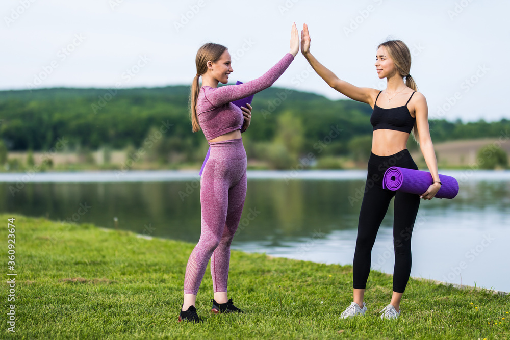 Two girls give each other high five after a good training session in the park near lake. Attractive sporty women smiling outdoor.