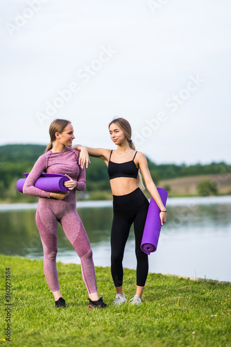 Two cheerful sportswomen in tracksuits talking and laughing while standing with yoga mat by lake in morning