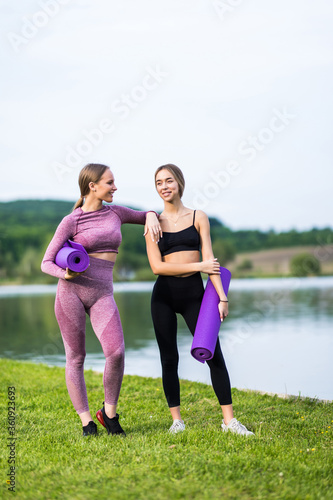 Two cheerful sportswomen in tracksuits talking and laughing while standing with yoga mat by lake in morning