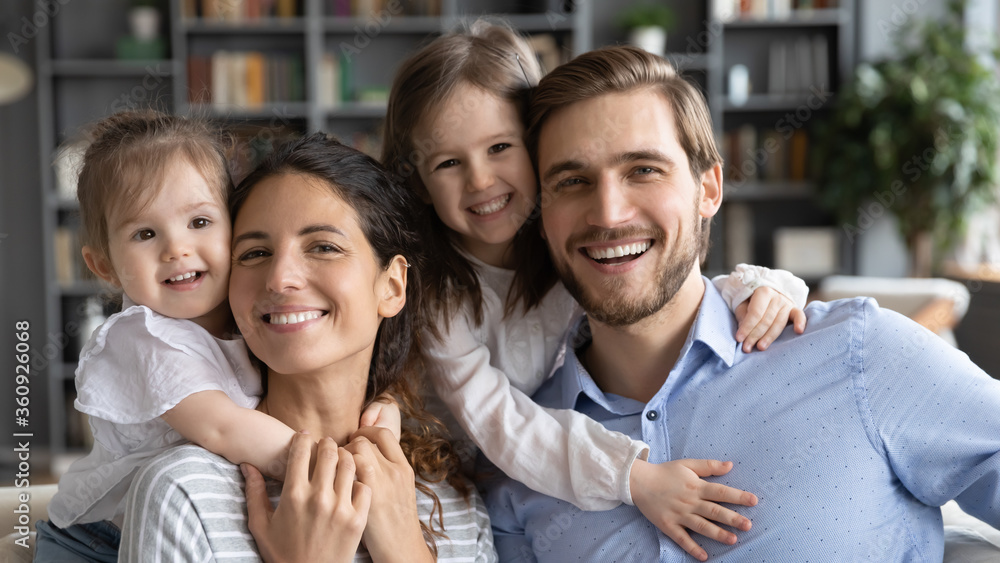 Obraz premium Head shot portrait happy young beautiful mother and smiling father piggy backing two laughing adorable little daughters, looking at camera, posing for family photo in modern living room at home