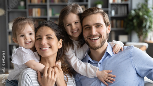Head shot portrait happy young beautiful mother and smiling father piggy backing two laughing adorable little daughters  looking at camera  posing for family photo in modern living room at home