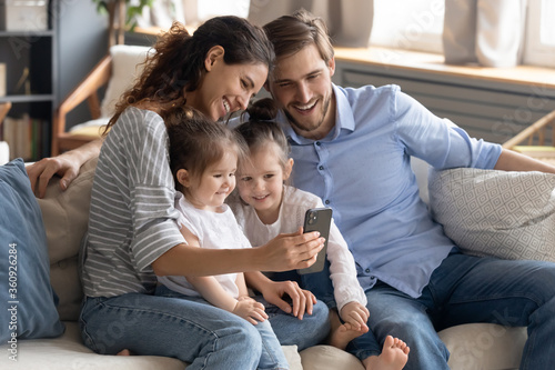 Smiling young mother and father with two adorable little daughters using smartphone, looking at screen, sitting on cozy couch at home, family making video call, watching cartoons, shopping online