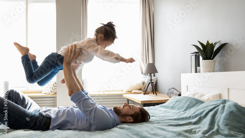 Smiling father holding little girl with hands outstretched pretending flying, relaxing in cozy bed in bedroom, happy young dad and adorable preschool daughter playing funny game in bedroom