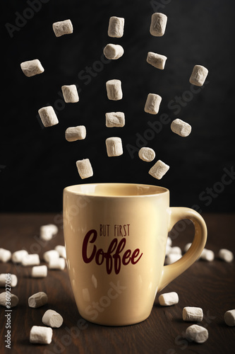 Beige coffee cup standing on wooden table with flying white marshmallows at kitchen against dark background