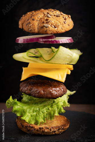 Flying layers of burger with beef meet, cheese, cucumber and onion rings against dark background