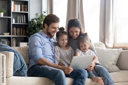 Happy young family with two little daughters using laptop together, smiling mother and father with adorable preschool girls looking at screen, watching cartoons, shopping online, making video call