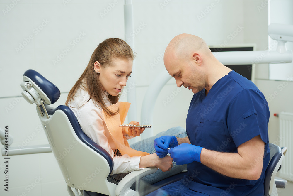 Obraz premium A bald middle-aged dentist shows a female patient tooth color samples before a whitening procedure in a dentist's office. A doctor is matching a tooth color to a patient before treatment.