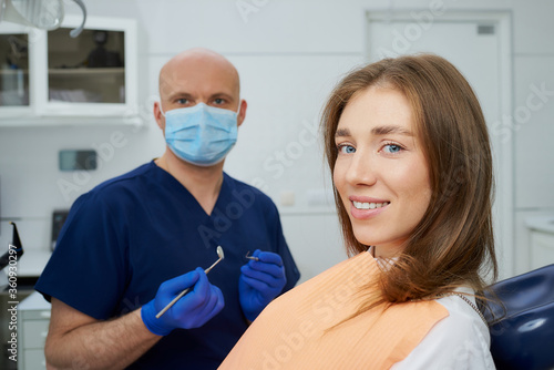A bald dentist in a medical face mask is holding a dental mirror and dental explorer near a cute patient in a dental chair. A doctor with a patient who is demonstrating a result of the tooth treatment