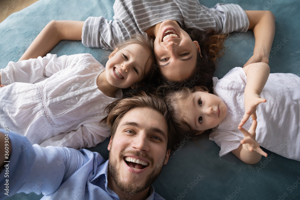 Top view head shot portrait close up happy young family lying on bed together, smiling beautiful mother and father with two cute little daughters looking at camera, posing for selfie, having fun Top view head shot portrait close up happy young family lying on bed together, smiling beautiful mother and father with two cute little daughters looking at camera, posing for selfie, having fun
