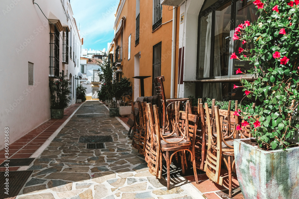 Outside view of a closed cafe during early morning in Spain. Bright sun making shadows on stacked chairs and colorful yellow and white buildings. Beautiful narrow Spanish street with red flowers.