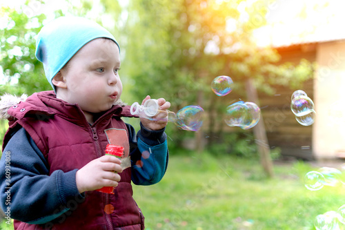 Cute little baby in spring blows soap bubbles. A boy on the street  dressed warmly. Bubbles are flying in different directions. The photo is artistically processed. horizontal