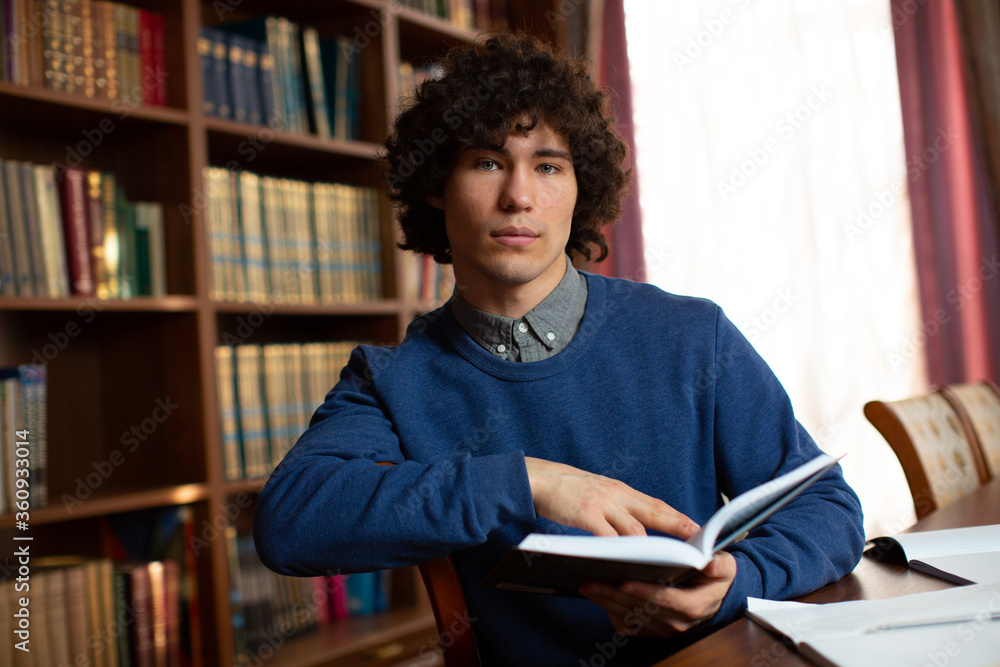 Portrait of a beautiful curly boy student in a gray shirt, and blue sweater with an open book against the background of bookshelves in the library Portrait of a beautiful curly boy student in a gray shirt, and blue sweater with an open book against the background of bookshelves in the library