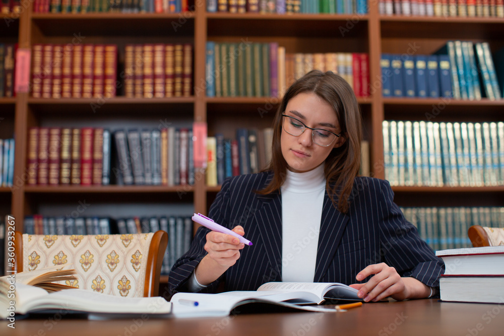 The girl student with dark hair and glasses in a gray striped jacket holds a pen in her hands, and focuses on the task in a notebook on a table against the background of a bookshelf The girl student with dark hair and glasses in a gray striped jacket holds a pen in her hands, and focuses on the task in a notebook on a table against the background of a bookshelf
