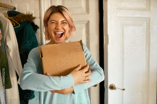Indoor image of happy cheerful young woman holding cardboard box delivered to her apartment, expressing excitement, going to unpack parcel, having impatient overjoyed look. Food delivery and shopping photo