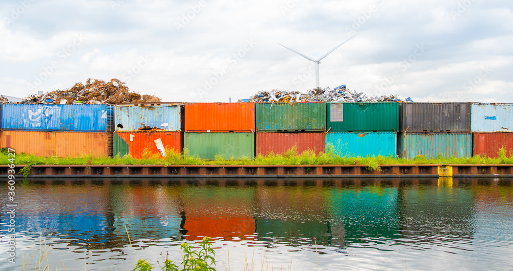 Metal scrap dump, with rusty metal parts on colorful, old truck containers, near water canal, river. Wind power windmills on background.