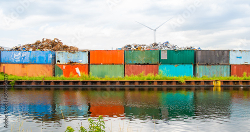 Obraz na płótnie Metal scrap dump, with rusty metal parts on colorful, old truck containers, near water canal, river. Wind power windmills on background. w salonie