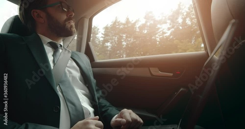 Thoughtful businessman in black suit and eyewear using portable computer while sitting on backseat of car. Busey bearded person working on laptop while driving. photo