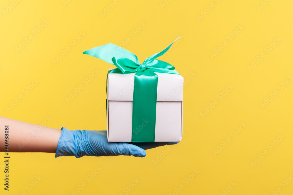 Profile side view closeup of human hand in blue surgical gloves holding white gift box. sharing, giving or delivery concept. indoor, studio shot, isolated on yellow background.