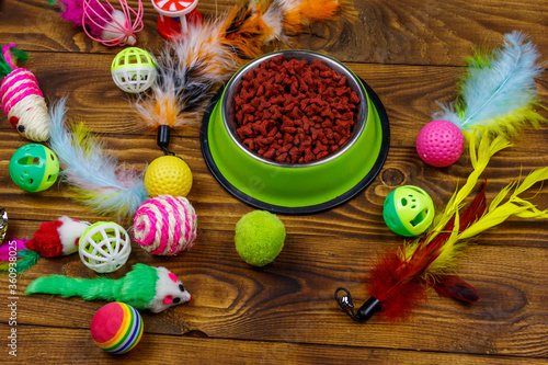 Set of toys for cat and bowl with dry pet food on wooden background. Pet care concept