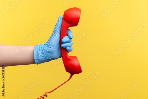 Profile side view closeup of human hand in blue surgical gloves holding and showing red call telephone handset receiver. indoor, studio shot, isolated on yellow background.