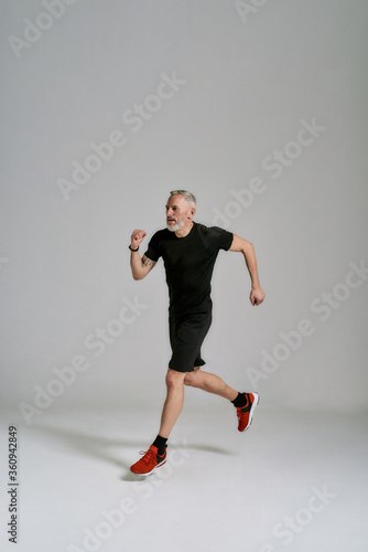 Full length shot of middle aged muscular man in black sportswear looking aside while jogging during workout in studio over grey background