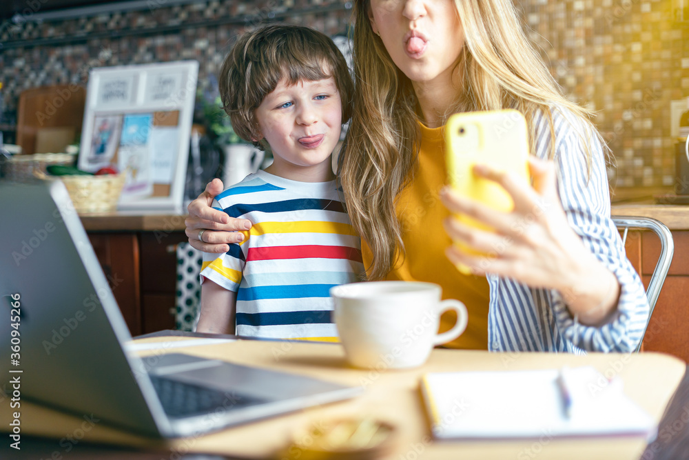 Mother and son show tongue, make selfie looking at smart phone. Having fun together at home. Woman working from home and online education with kid with computer and phone Mother and son show tongue, make selfie looking at smart phone. Having fun together at home. Woman working from home and online education with kid with computer and phone