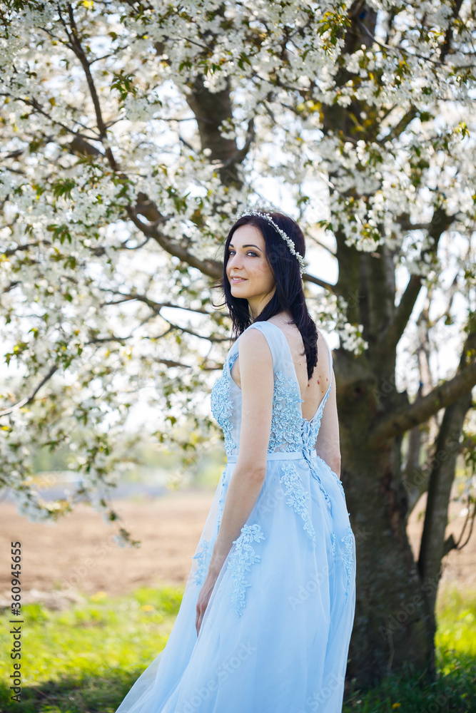 Beautiful girl in a light summer blue long dress adorned in her hair against a blossoming tree. Tender portrait of a young woman in white blossom
