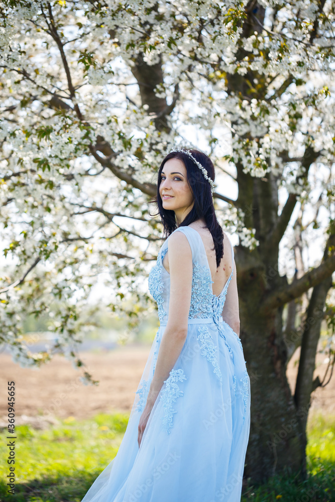 Beautiful girl in a light summer blue long dress adorned in her hair against a blossoming tree. Tender portrait of a young woman in white blossom Beautiful girl in a light summer blue long dress adorned in her hair against a blossoming tree. Tender portrait of a young woman in white blossom