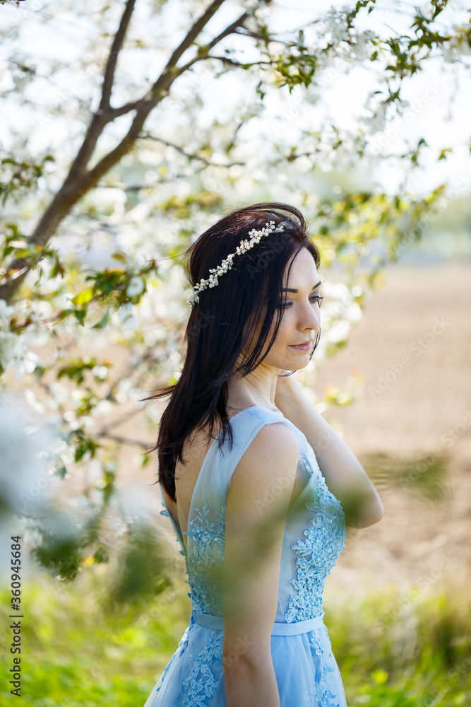 Obraz premium Beautiful girl in a light summer blue long dress adorned in her hair against a blossoming tree. Tender portrait of a young woman in white blossom