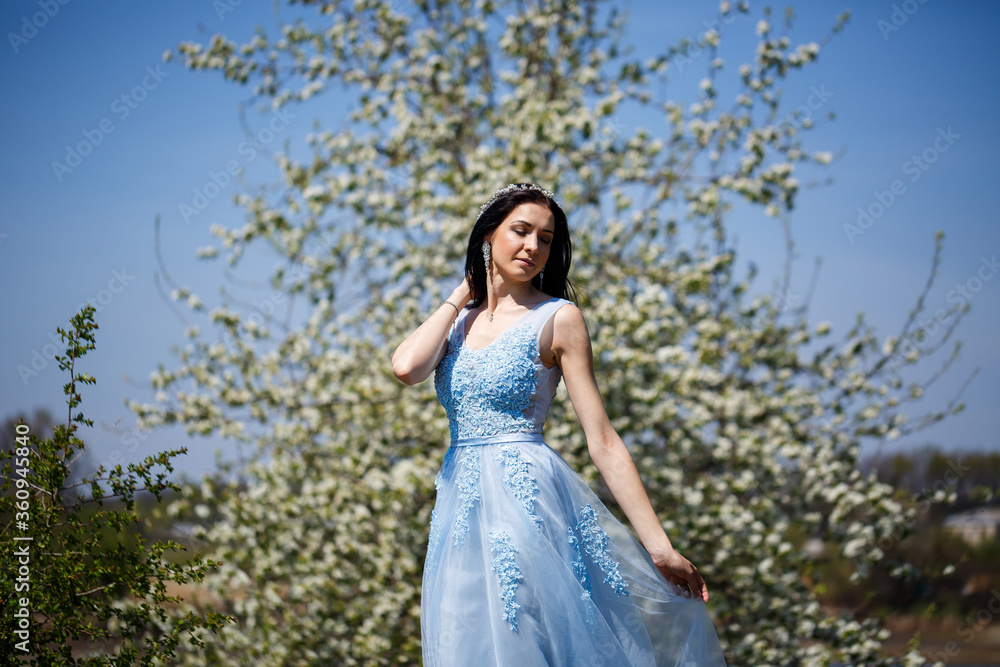 Young woman with beautiful make-up with a vintage hoop in a blue light dress with a bouquet posing on a background of green foliage in a park. Fresh portrait attractive girl with white flowers. Young woman with beautiful make-up with a vintage hoop in a blue light dress with a bouquet posing on a background of green foliage in a park. Fresh portrait attractive girl with white flowers.