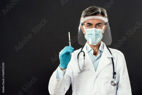 Mature doctor in medical mask and face shield holding a syringe with vaccine from flu, coronavirus, covid-19 while standing against black background