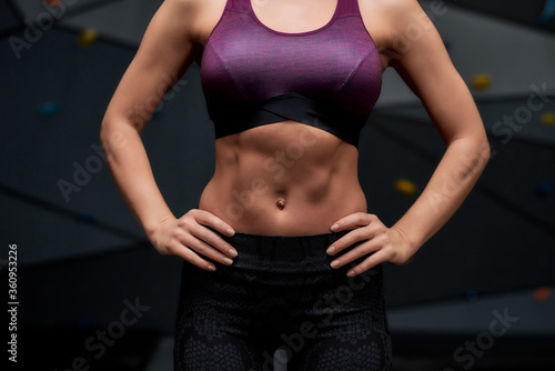 Cropped shot of active young woman in sportswear showing her sportive body, abs, standing against artificial training climbing wall. Concept of sport life and rock climbing