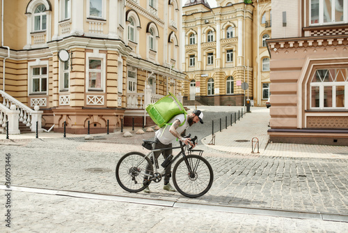 Something wrong. Full length shot of brutal bearded delivery man in cap checking his bicycle, while riding it outdoors. Courier, delivery service concept