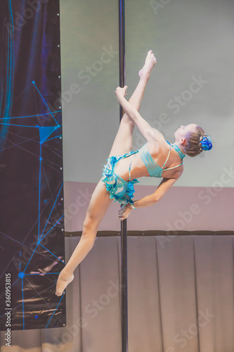 Picture of girl in light blue costume is showing acrobatic ticks on pylon. Straight legs above the head, as activity during quarantine. Little girl on championship on pole dance art.