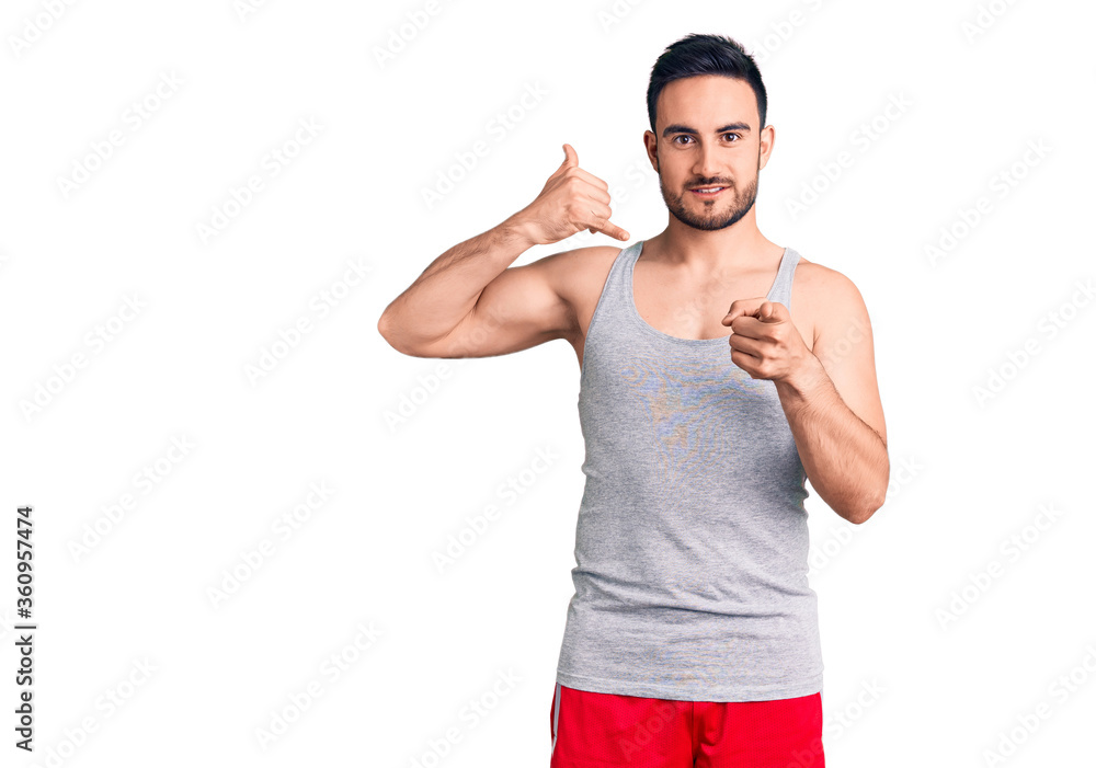 Young handsome man wearing swimwear and sleeveless t-shirt smiling doing talking on the telephone gesture and pointing to you. call me. Young handsome man wearing swimwear and sleeveless t-shirt smiling doing talking on the telephone gesture and pointing to you. call me.