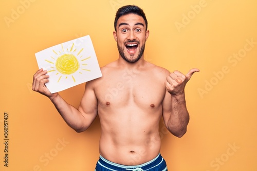 Young handsome man with beard on vacation wearing swimwear holding paper with sun draw pointing thumb up to the side smiling happy with open mouth