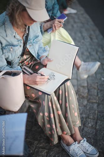 Young beautiful girl draws in a park in a group. Outline in a sketchbook.