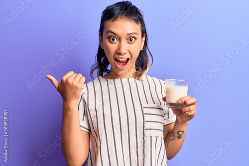 Young woman holding glass of milk pointing thumb up to the side smiling happy with open mouth